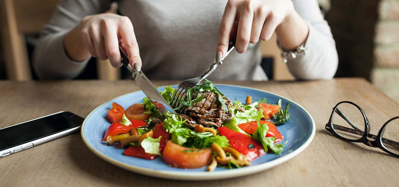 Fotografia de uma pessoa utilizando talheres para se alimentar de verduras, legumes e carne em um prato.