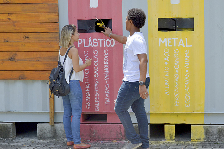 Fotografia de duas pessoas em frente a um ponto de coleta seletiva de lixo, descartando materiais.