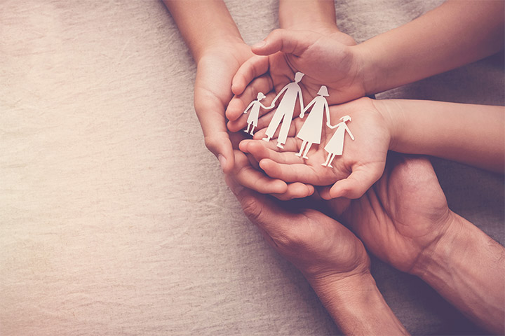 Fotografia de três pessoas sobrepondo suas mãos, em que carregam uma representação em recorte de papel de uma família tradicionalmente composta por um pai, uma mãe e duas crianças.