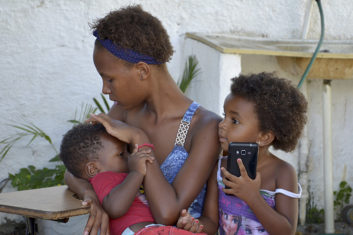 Fotografia em que aparece uma mulher amamentando um bebê enquanto outra criança está ao lado, segurando um aparelho celular.