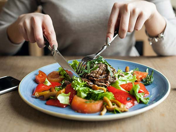 Fotografia de uma pessoa utilizando talheres para se alimentar de verduras, legumes e carne em um prato.