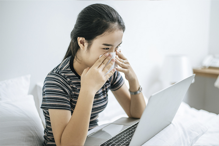 Fotografia de uma mulher em frente à tela de um computador laptop assoando o nariz.