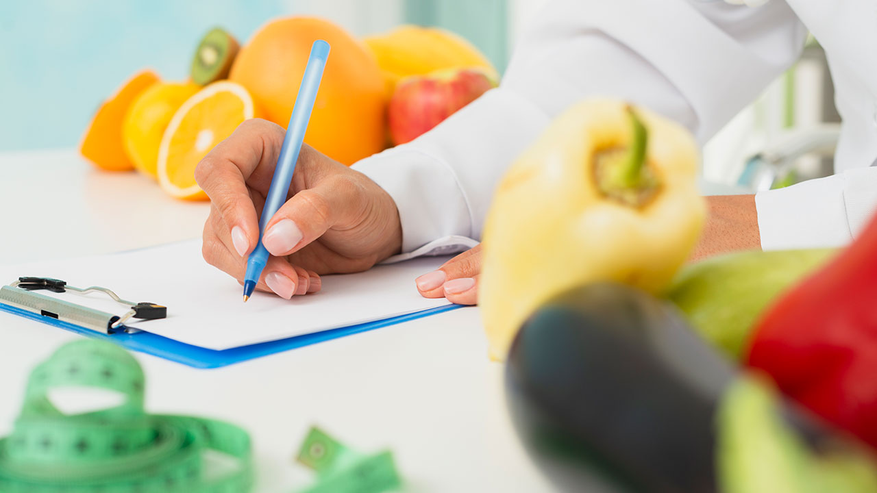 Foto de uma pessoa de jaleco branco escrevendo com uma caneta em um papel preso a uma prancheta em cima de uma mesa branca, o foco da foto está nas mãos, ao fundo há laranjas, maçã e outras frutas, em primeiro plano da foto e desfocado, uma fita métrica e alguns pimentões.