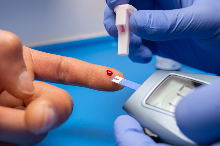 Foto de um medidor de glicose sendo segurado por mãos com luvas azuis, ao lado um dedo com uma gota de sangue.