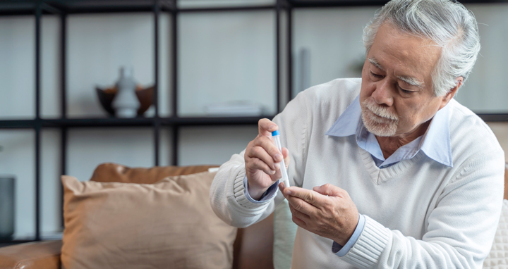 Foto de um senhor branco, com barba e cabelos brancos, ele veste uma blusa branca de mangas compridas com uma blusa azul de gola embaixo, ele está sentado em um sofá, segurando um pequeno tubo com uma mão enquanto coloca a base do tubo no dedo da outra mão.