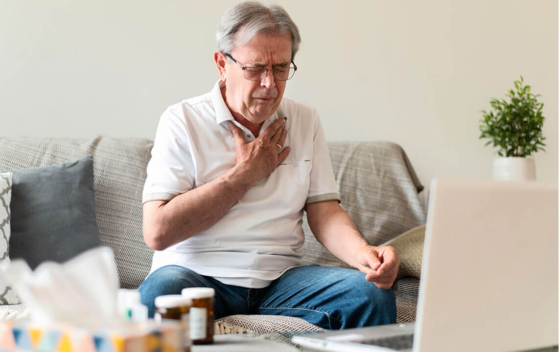 Foto de um senhor branco com cabelos grisalhos, ele veste uma blusa de manga curta com gola branca, calça jeans e óculos, está sentado no sofá, na frente dele há um laptop e alguns remédios, está com a mão próxima ao pescoço, pela expressão em seu rosto está sentindo dor.