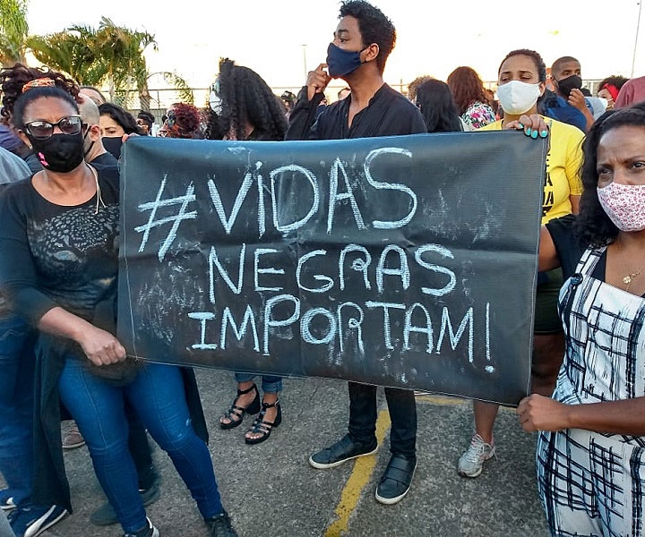 Fotografia de uma manifestação com diversas pessoas, a maioria pessoas negras, utilizando máscaras faciais. Em primeiro plano, duas mulheres negras utilizando máscaras faciais, segurando um cartaz escrito “#Vidas negras importam!”