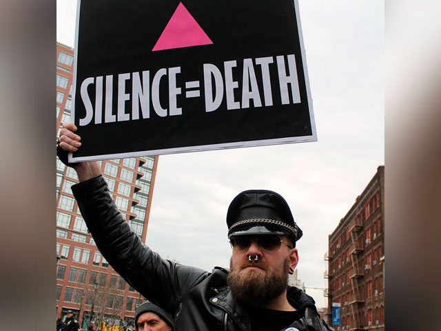 Fotografia de um homem branco, com barba comprida, usando óculos escuros, boina preta, casaco preto e piercing no nariz. Esse homem segura um cartaz com dizeres em inglês “Silence = Death”, tradução livre para português “Silêncio é igual à morte”.