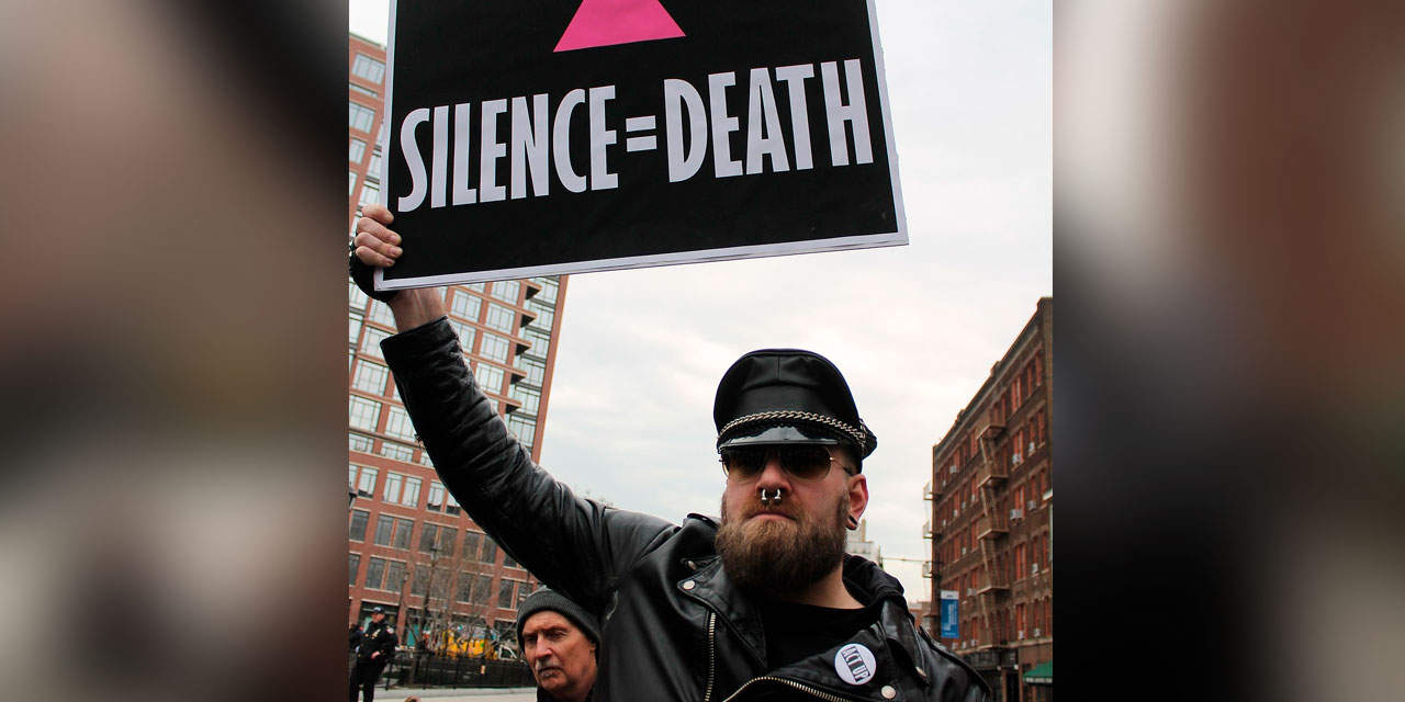 Fotografia de um homem branco, com barba comprida, usando óculos escuros, boina preta, casaco preto e piercing no nariz. Esse homem segura um cartaz com dizeres em inglês “Silence = Death”, tradução livre para português “Silêncio é igual à morte”.