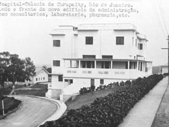 Fotografia em preto e branco do edifício do Hospital colônia de Curupaity. O edifício é uma casa branca, com janelas quadradas em três níveis/ andares com um espaço de uma varanda que contorna todo o edifício, localizado em meio a jardins e ruas arborizadas.