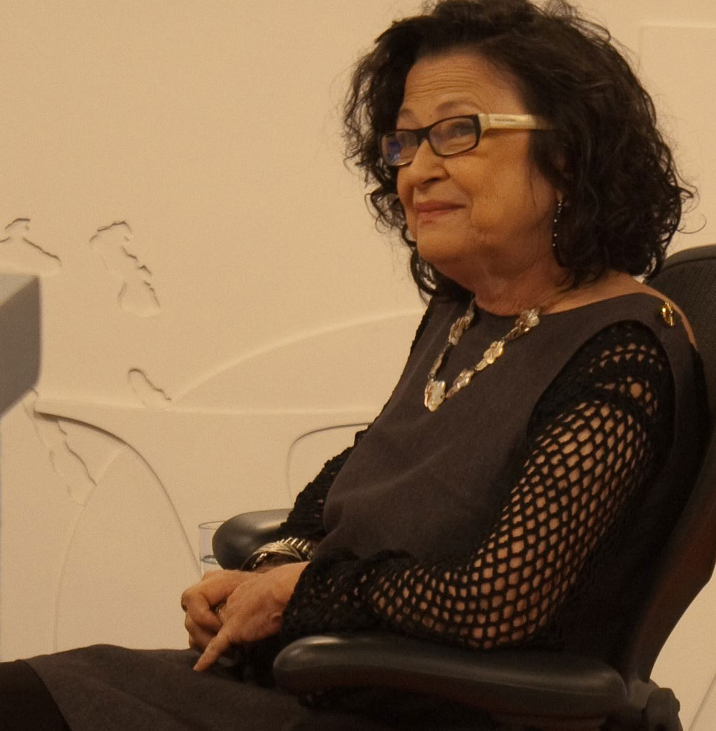 Fotografia colorida quadrada mostrando uma mulher sentada em uma cadeira de auditório preta. Ela veste vestido preto de mangas longas, colares de pedras e usa óculos. Os cabelos são castanhos e cacheados, na altura dos ombros. A fotografia só mostra a parte superior do corpo sentado.