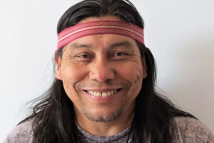 Fotografia do indígena Daniel Mundukuru, em que olha para que faz a foto e sorri. Ele utiliza um adereço vermelho (faixa) na cabeça, tem cabelos longos e pretos abaixo dos ombros e está vestindo uma camiseta cinza.