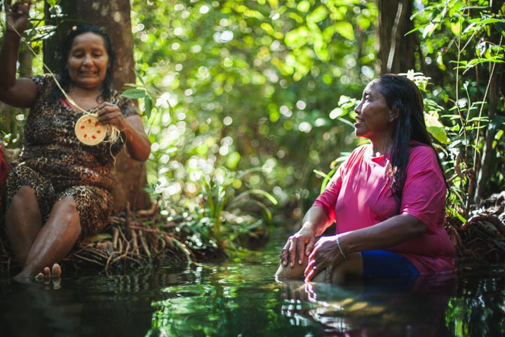 Fotografia de duas mulheres indígenas sentadas em meio a uma região de floresta. Elas conversam entre si, enquanto uma delas faz um trabalho de artesanato. Uma delas usa um vestido colorido e a outra uma camiseta rosa.