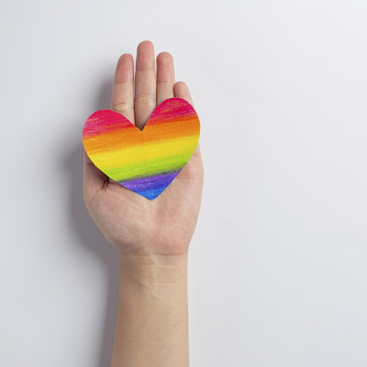 Fotografia de uma mão de uma pessoa branca, com o desenho de um coração de papel com as cores na horizontal, formando um arco-íris  (vermelho, laranja, amarelo, verde, roxo e azul)