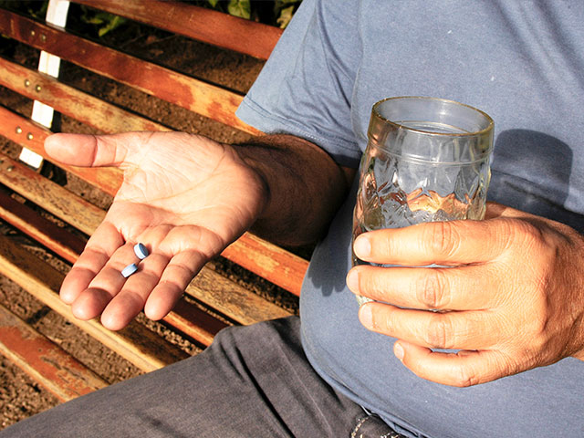 Fotografia de uma pessoa branca, que em uma mão segura dois comprimidos azuis e na outra mão, um copo de vidro contendo água.
