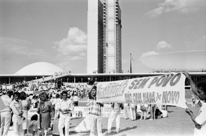 Foto em preto e branco, há diversas pessoas no Palácio do Planalto em Brasília subindo a rampa e no gramado, em primeiro plano na foto algumas pessoas seguram faixas, é possível ler em uma delas: “Constituinte, sem povo não cria nada de novo”