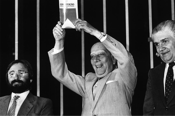 Foto em preto e branco de três homens usando terno, no centro está o presidente da Assembleia Nacional Constituinte Ulysses Guimarães erguendo a constituição, ao seu lado os deputados Marcelo Cordeiro e Mauro Benevides.