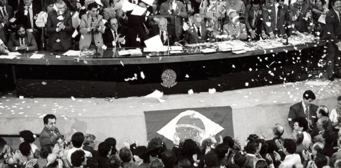 Foto em preto e branco da câmara dos deputados. No plenário tem diversas pessoas em pé e sentadas, em frente ao plenário há uma bandeira do Brasil e pessoas em pé, está voando papel branco.