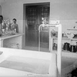 fotografia em preto e branco da sala de meios de cultura no Castelo. na frente da imagem, bastante desfocada, está a torneira de um aparato hidráulico, inventado por um dos cientistas do IOC, para produção de água destilada. ao fundo, uma bancada de azulejos com a vidraria e atrás da bancada, dois trabalhadores auxiliares em destaque.