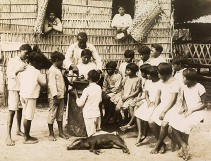 Foto nas cores preto e branco. No centro, crianças indígenas Parintintins ouvem um gramofone. Algumas crianças estão de pé, outras sentadas, observando a presença de um homem negro, que manuseia o instrumento. Também na imagem se observa um animal deitado no chão, próximo às crianças. Ao fundo, duas crianças indígenas observam a cena da janela de suas casas.