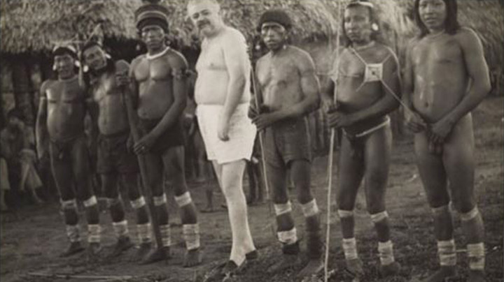 Foto nas cores preto e branco. Há seis indígenas, posicionados de frente, três de cada lado, e, ao centro, um homem branco, sem camisa, também posicionado de frente.