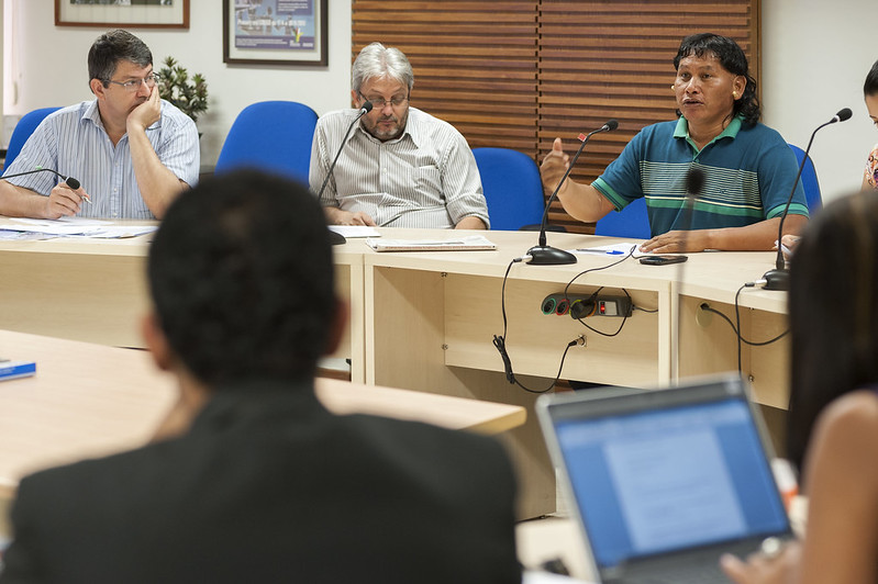 Foto da “Nonagésima primeira reunião da Comissão Intersetorial de Saúde Indígena”. Há a presença de um indígena, vestindo uma camisa azul e verde. Ele está sentando numa bancada, ao lado de outras pessoas, falando a um microfone. No primeiro plano, há a presença de pessoas  que o observam falar, e uma pessoa que escreve num laptop.