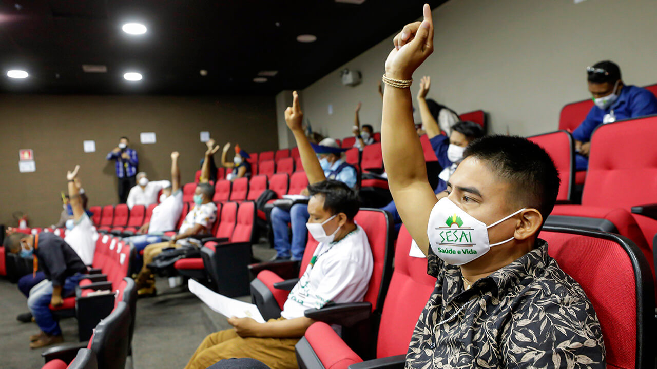 Foto do Fórum de presidentes do Conselho Distrital de Saúde Indígena. Na imagem, a presença de indígenas em um auditório com cadeiras vermelhas. Todos usam máscaras de proteção facial. Alguns levantam os braços em sinal de representação de seus votos.