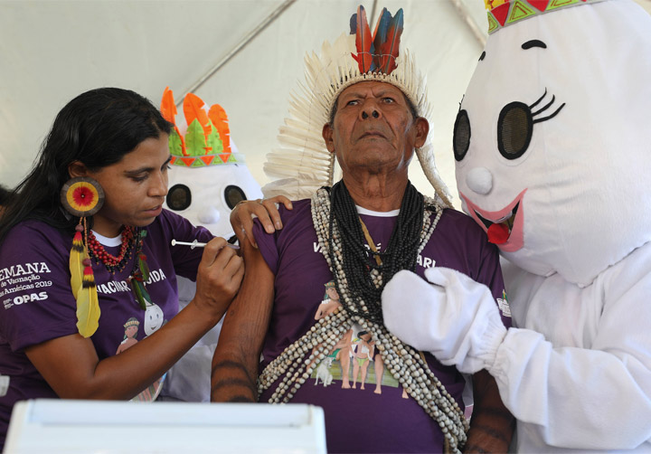 Foto de um indígena recebendo a dose de uma vacina, aplicada por uma profissional de saúde também indígena. Ambos vestem uma camisa roxa e usam adereços típicos. O indígena recebe um abraço do mascote da campanha de vacinação.