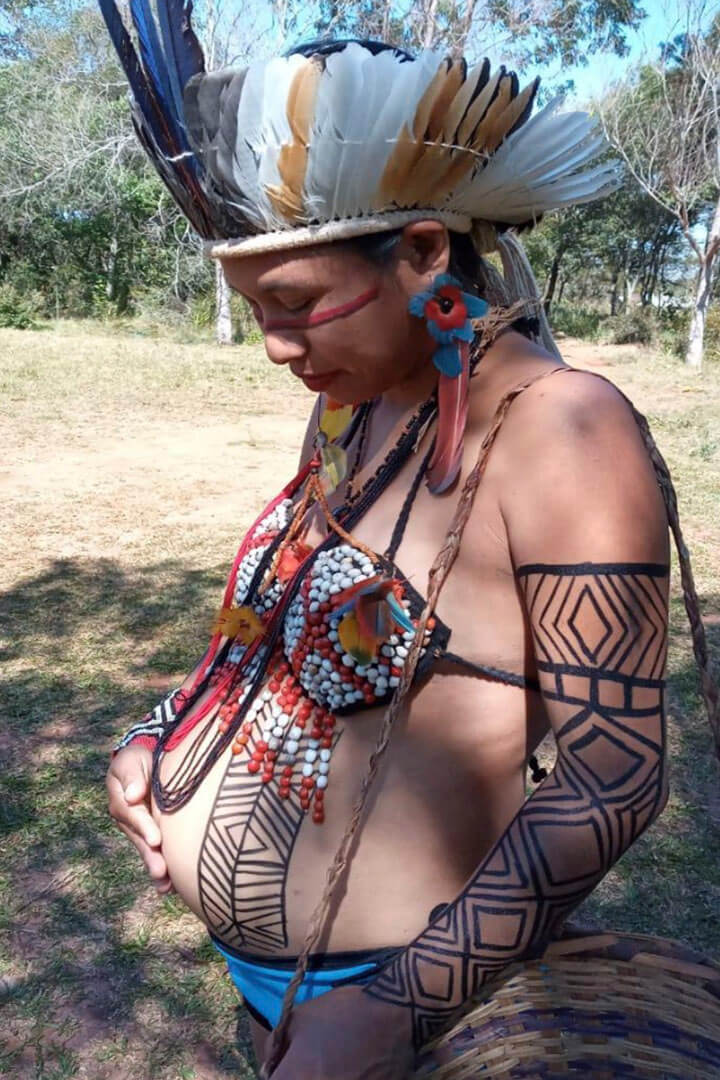 Foto de uma gestante indígena do povo Guarani. Ela usa adereços e pintura corporal típicos, e toca a barriga com uma das mãos.