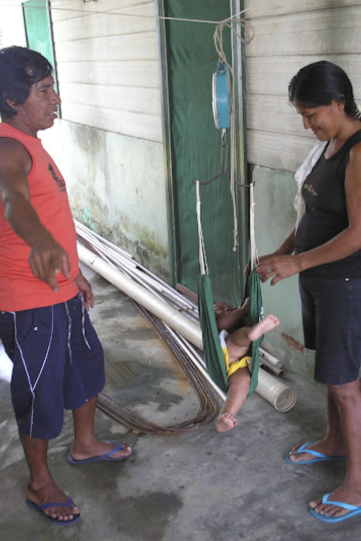 Foto de um agente indígena de saúde na aldeia Kwatá, Amazonas. O agente realiza a pesagem de um bebê enquanto conversa com sua mãe.