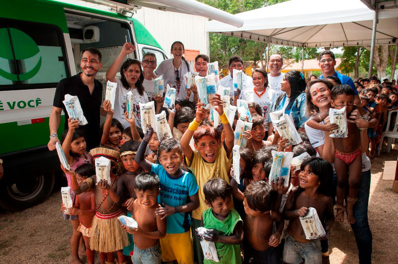 Foto da equipe do Programa “Brasil Sorridente” onde há a presença de diversas crianças indígenas sorrindo entre os profissionais de saúde. As crianças seguram kits contendo material de higiene dental.