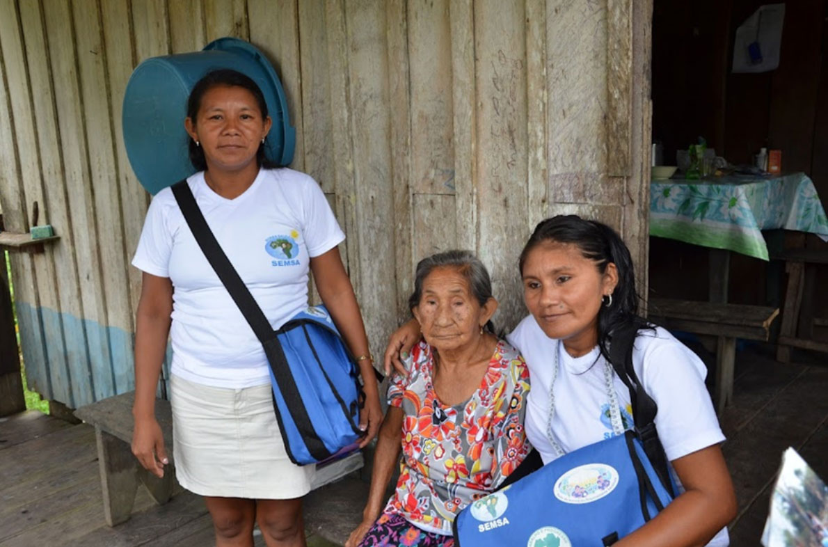 Foto de duas agentes indígenas de saúde Munduruku, fazendo visita domiciliar na aldeia Kwatá, Amazonas. Elas vestem camisas brancas e abraçam uma idosa indígena que está sentada em uma cadeira do lado de fora de sua casa.