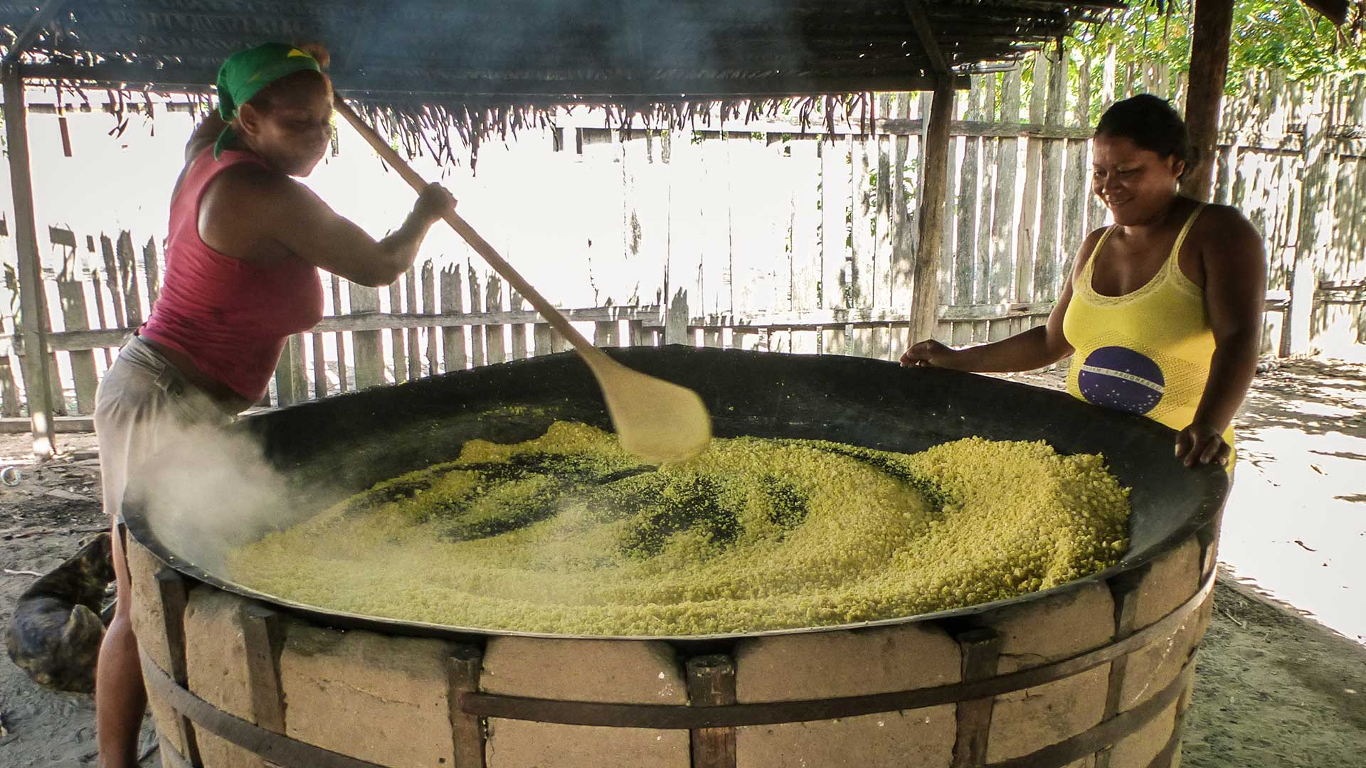 Fotografia de duas mulheres de comunidades tradicionais, elas estão em frente a uma panela enorme, uma de cada lado, a que está do lado esquerdo veste uma blusa rosa e segura uma grande colher de madeira, enquanto mistura o conteúdo na panela, a mulher do lado direito veste uma blusa do Brasil e sorri enquanto olha para o centro da panela, ao fundo há um muro feito com estacas de madeira.