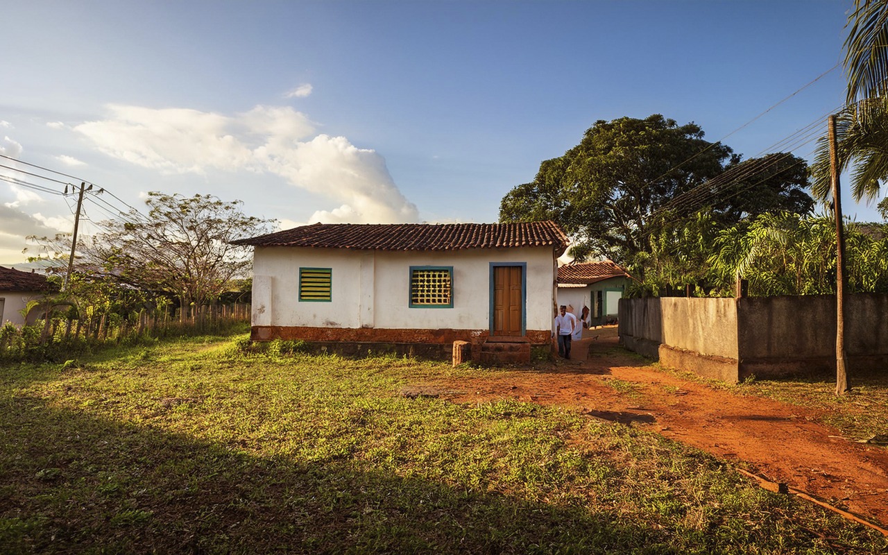 Imagem de uma casa branca, com duas janelas e uma porta de madeira. Há pessoas do lado direito da casa, na frente da casa há grama aparada, do lado esquerdo há um poste de energia e árvores, do lado direito há fios de energia, árvores e um muro.
