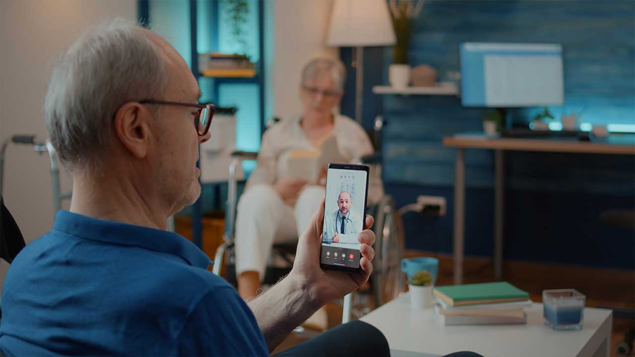 Fonte: Freepik Fotografia de um homem branco, idoso, de óculos segurando um celular em videoconferência com um médico.