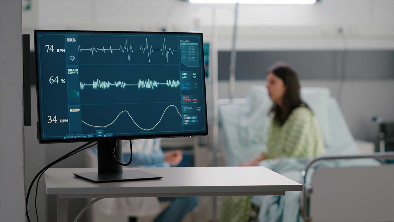 Fonte: Freepik Fotografia de um monitor médico com batimentos cardíacos, ao fundo uma mulher sentada em uma cama de hospital.